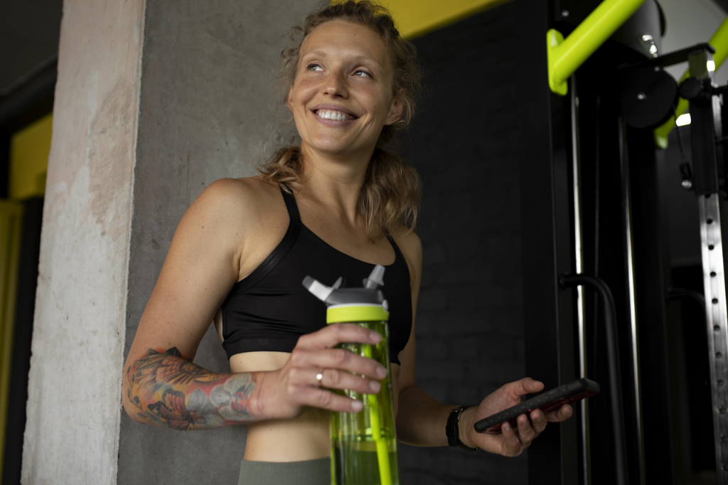 A woman enjoying her gym class while drinking a pre workout supplement shake, with gym equipment visible in the background
